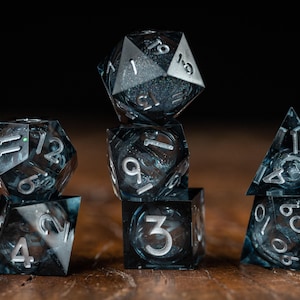 May include: A collection of six polyhedral dice, ideal for role-playing games. The dice are black with silver numerals. They are displayed on a wooden surface against a dark backdrop.