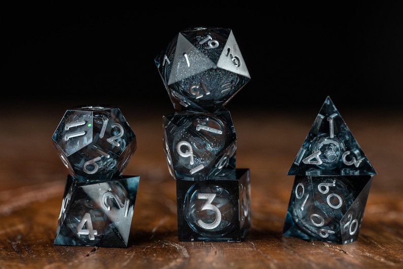May include: A collection of six polyhedral dice, ideal for role-playing games. The dice are black with silver numerals. They are displayed on a wooden surface against a dark backdrop.