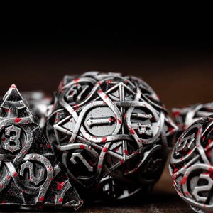May include: Close-up of a set of metallic dice with intricate designs and red accents. The dice include a pyramid, a sphere, and other geometric shapes, ideal for tabletop gaming. The numbers are clearly visible.