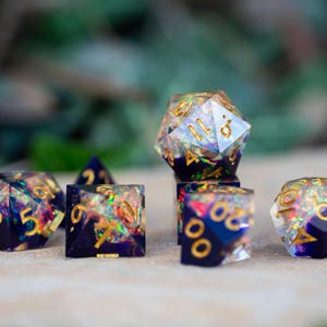 May include: A set of seven polyhedral dice with a dark blue base and gold numbering. The dice are filled with iridescent glitter, creating a colorful, shimmering effect. The dice are for tabletop gaming.
