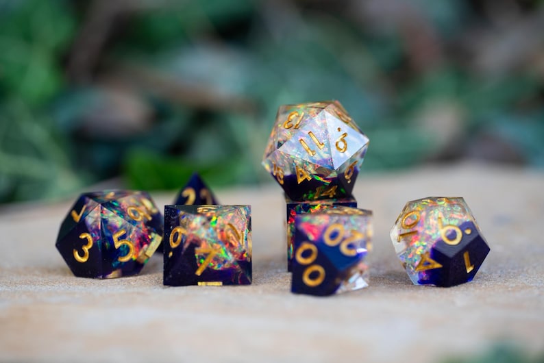 May include: A set of seven polyhedral dice with a dark blue base and gold numbering. The dice are filled with iridescent glitter, creating a colorful, shimmering effect. The dice are for tabletop gaming.