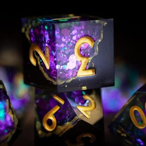May include: Close-up of several polyhedral dice with a black base, filled with purple, blue, and gold glitter. The numbers are gold. The dice are for tabletop role-playing games.