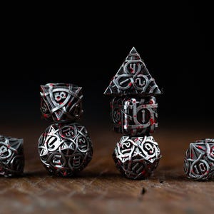 May include: A collection of intricately designed dice, featuring a silver metallic finish with red accents. The dice are arranged on a wooden surface against a dark background, showcasing various shapes and sizes, ideal for tabletop gaming.