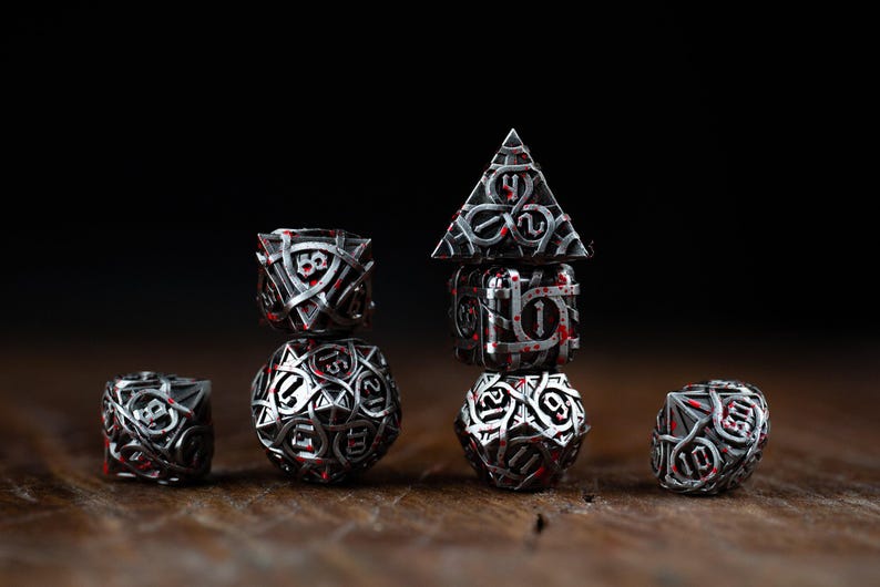 May include: A collection of intricately designed dice, featuring a silver metallic finish with red accents. The dice are arranged on a wooden surface against a dark background, showcasing various shapes and sizes, ideal for tabletop gaming.