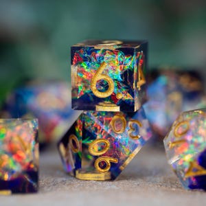 May include: A set of handcrafted resin dice with a dark blue base and colorful, iridescent inclusions. The dice feature gold-colored numbers and are designed for tabletop gaming.