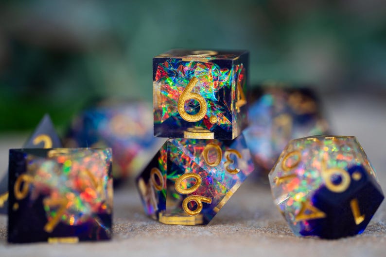 May include: A set of handcrafted resin dice with a dark blue base and colorful, iridescent inclusions. The dice feature gold-colored numbers and are designed for tabletop gaming.