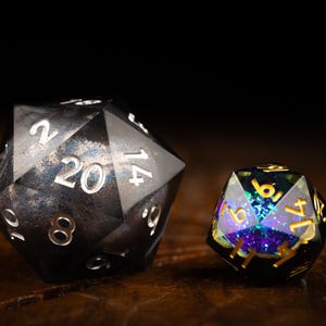 Puede incluir: Dos dados poliédricos, uno grande y negro con números blancos, el otro más pequeño con un interior brillante y colorido y números dorados. Los dados son para juegos de rol de mesa.