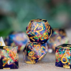 May include: A collection of polyhedral dice with a dark blue base and gold numbering. The dice are filled with colorful, iridescent glitter, creating a shimmering effect. The set includes various shapes, such as a d20, d6, and d4, perfect for tabletop gaming.