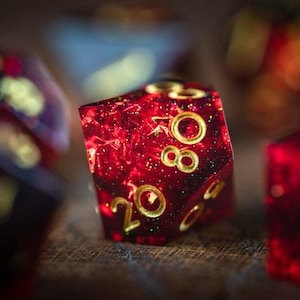 Pode incluir: Close-up de um dado vermelho de vinte lados cheio de glitter, com numeração dourada. Outros dados estão desfocados no fundo. Os dados são para jogos de tabuleiro.