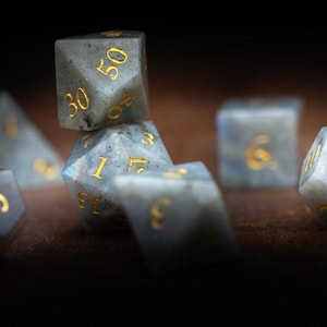 Puede incluir: Un conjunto de dados de piedras preciosas para juegos de rol. Los dados tienen varias formas, incluyendo un d20, d12 y d6, con n&uacute;meros dorados. Los dados son una mezcla de gris y azul con un patr&oacute;n moteado.