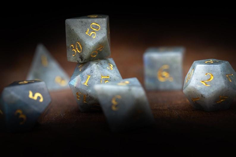 Puede incluir: Un conjunto de dados de piedras preciosas para juegos de rol. Los dados tienen varias formas, incluyendo un d20, d12 y d6, con n&uacute;meros dorados. Los dados son una mezcla de gris y azul con un patr&oacute;n moteado.