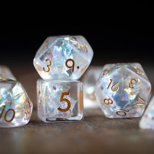 May include: A collection of clear, iridescent dice with gold-colored numbers. The dice are various shapes, including a six-sided die, a ten-sided die, and a twenty-sided die. The dice are arranged on a wooden surface.