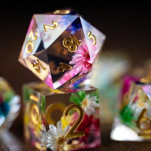 May include: Close-up of several clear resin dice with gold numbering and colorful floral inclusions. The dice are in various shapes, including a twenty-sided die and a six-sided die. The flowers are pink, white, and yellow.