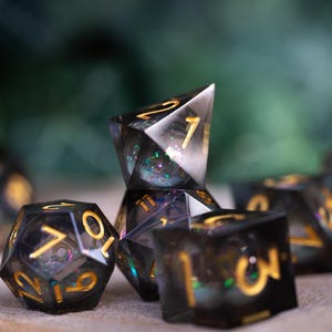 May include: A set of seven black and gold iridescent dice with a variety of shapes and numbers. The dice are made of clear resin with a glittery finish.