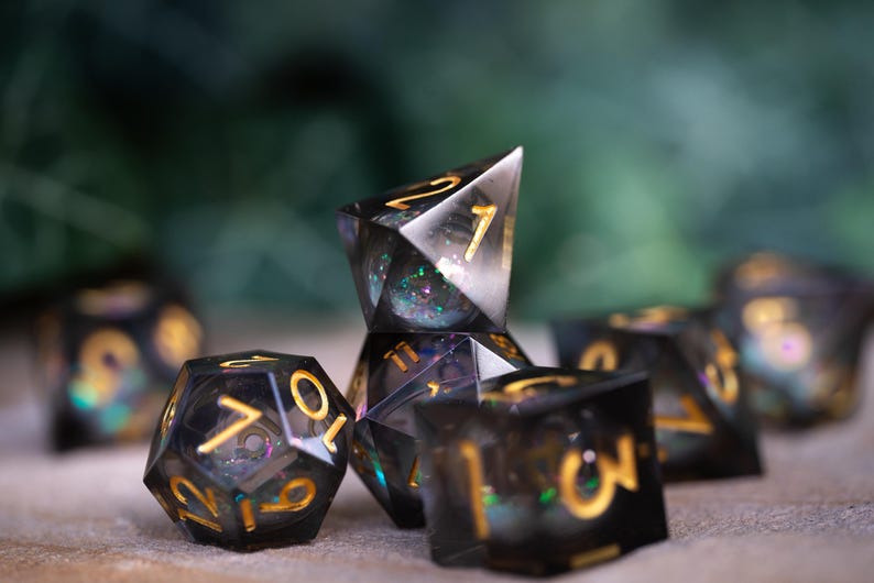 May include: A set of seven black and gold iridescent dice with a variety of shapes and numbers. The dice are made of clear resin with a glittery finish.