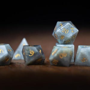 Puede incluir: Un conjunto de dados de piedras preciosas en varias formas, incluyendo un d20, d10, d6 y d4. Los dados son una mezcla de gris y azul con n&uacute;meros dorados. Los dados est&aacute;n dispuestos sobre una superficie de madera oscura.