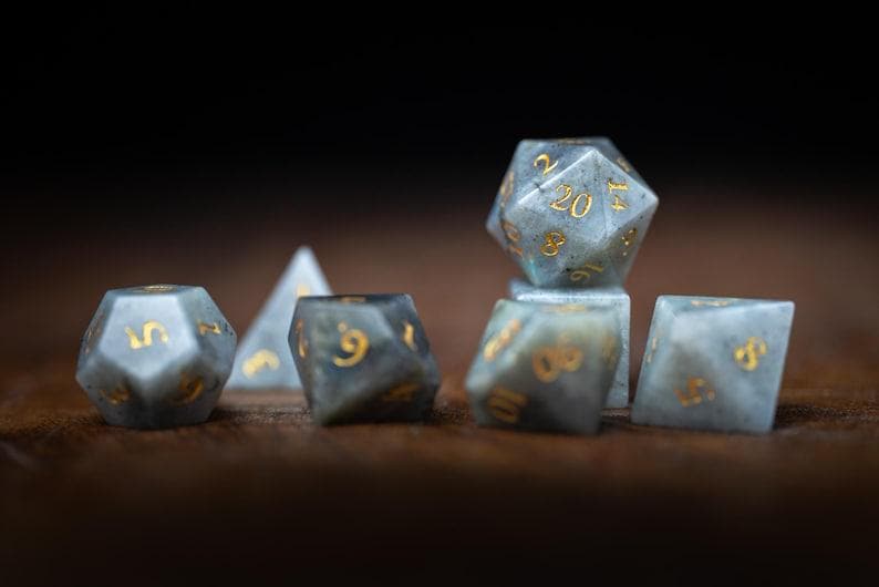 Puede incluir: Un conjunto de dados de piedras preciosas en varias formas, incluyendo un d20, d10, d6 y d4. Los dados son una mezcla de gris y azul con n&uacute;meros dorados. Los dados est&aacute;n dispuestos sobre una superficie de madera oscura.