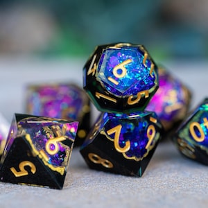 Könnte beinhalten: Eine Sammlung von Würfeln mit einem Galaxien-Design. Die Würfel sind schwarz mit goldenen Zahlen und gefüllt mit blauem und lila Glitzer. Geeignet für Tischrollenspiele.