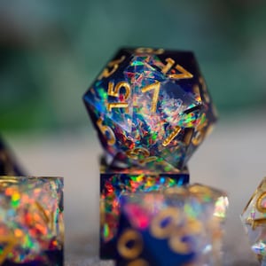 May include: Close-up of a set of iridescent resin dice. The dice are a deep blue with gold numbering and embedded with colorful glitter. The focus is on a twenty-sided die, with other dice in the background.