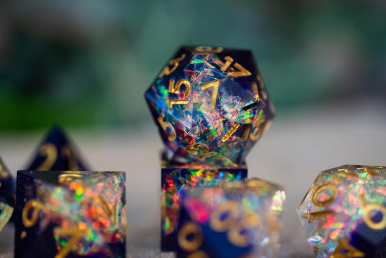May include: Close-up of a set of iridescent resin dice. The dice are a deep blue with gold numbering and embedded with colorful glitter. The focus is on a twenty-sided die, with other dice in the background.