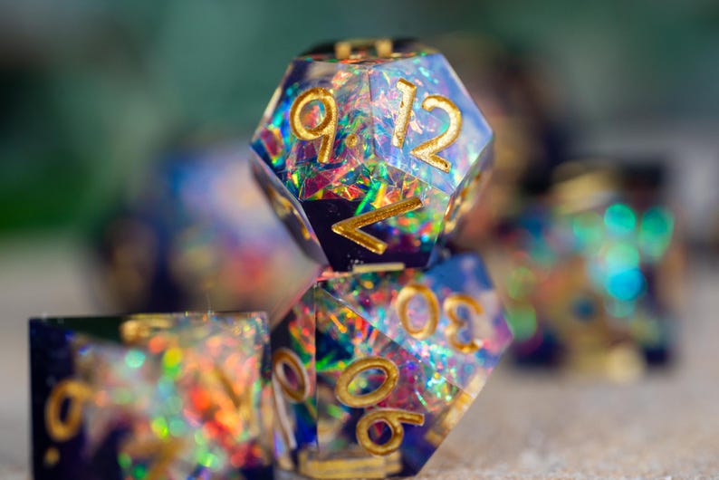 May include: Close-up of several polyhedral dice with a shimmering, iridescent interior. The dice have gold-colored numbers and edges. The dice are primarily blue with colorful glitter and are designed for tabletop gaming.
