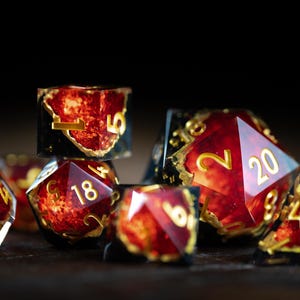 May include: A set of polyhedral dice featuring a translucent red and black resin design with gold-colored numbers. The dice have sharp edges and a swirling, fiery appearance within the material, with one die clearly showing the number 20.