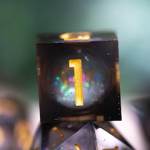 May include: A black and clear resin dice with gold glitter and a gold number "1" on one side.