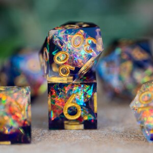 May include: Close-up of a set of iridescent resin dice with gold numbering. The dice are a mix of square and multi-sided shapes, with a dark blue base and colorful, shimmering inclusions. The numbers are in gold.