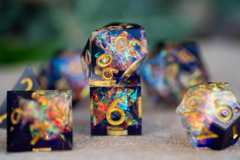 May include: Close-up of a set of iridescent resin dice with gold numbering. The dice are a mix of square and multi-sided shapes, with a dark blue base and colorful, shimmering inclusions. The numbers are in gold.