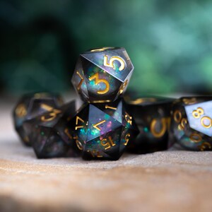 May include: A set of black and iridescent dice with gold numbers. The dice are made of resin and have a speckled appearance. The dice are arranged in a pyramid shape.