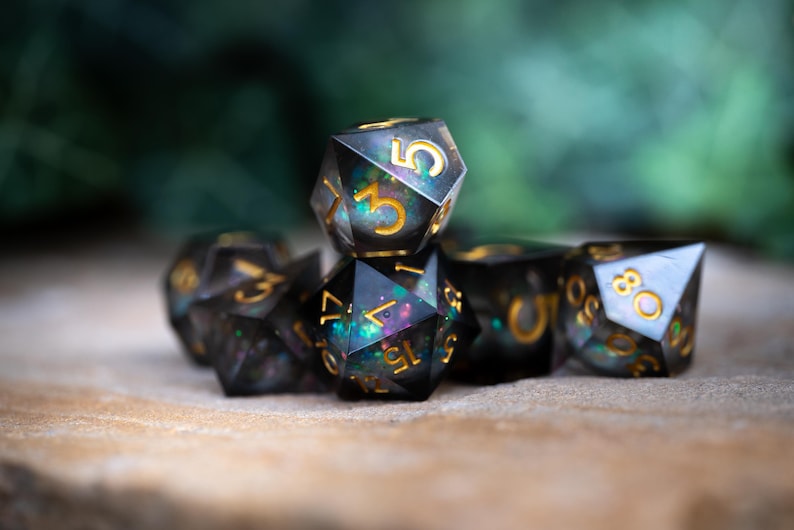 May include: A set of black and iridescent dice with gold numbers. The dice are made of resin and have a speckled appearance. The dice are arranged in a pyramid shape.