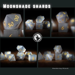 Puede incluir: Un conjunto de dados Moonshade Shards. Los dados son de color gris transl&uacute;cido con n&uacute;meros dorados. La imagen incluye varias formas de dados, incluyendo D20, D6 y D4. El texto "MOONSHADE SHARDS" est&aacute; en la parte superior.