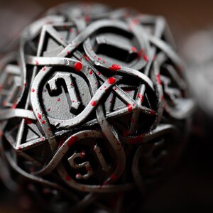 May include: Close-up of a metallic D20 die with intricate Celtic knot-like designs. The die is silver with red splatters, and the number 1 is visible. This dice is for tabletop gaming.