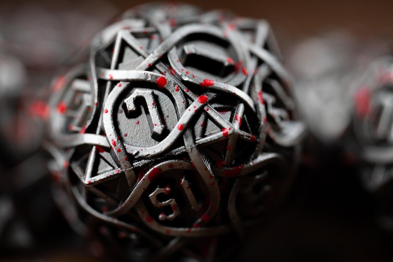 May include: Close-up of a metallic D20 die with intricate Celtic knot-like designs. The die is silver with red splatters, and the number 1 is visible. This dice is for tabletop gaming.