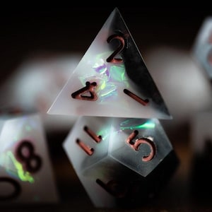 Peut inclure: Gros plan sur un ensemble de dés polyédriques. Les dés sont blancs avec des paillettes irisées et des chiffres de couleur cuivre. Les dés sont de formes variées, dont une pyramide et un dé à plusieurs faces.