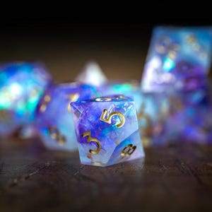 May include: A set of iridescent dice with gold numbering. The dice are a mix of blue, purple, and white, with a shimmering, galaxy-like appearance. The dice are designed for tabletop gaming.