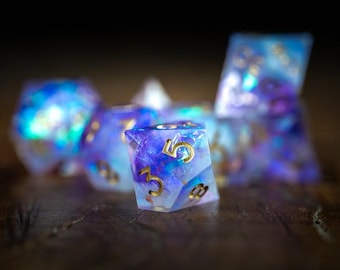 May include: A set of iridescent dice with gold numbering. The dice are a mix of blue, purple, and white, with a shimmering, galaxy-like appearance. The dice are designed for tabletop gaming.