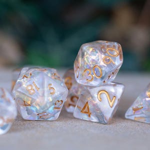 May include: A collection of iridescent, clear dice with gold-colored numbering. The dice are in various polyhedral shapes, including a d20, d12, d10, d8, d6, and d4. The dice are designed for tabletop role-playing games.