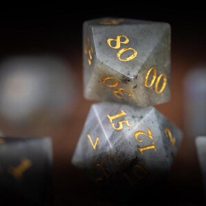 Puede incluir: Primer plano de un juego de dados de piedras preciosas. Los dados son grises con n&uacute;meros dorados. Los dados est&aacute;n apilados uno encima del otro, con el dado superior mostrando el n&uacute;mero 80. Los dados son para juegos de mesa.