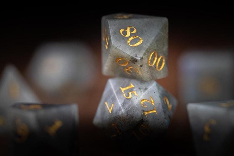 Puede incluir: Primer plano de un juego de dados de piedras preciosas. Los dados son grises con n&uacute;meros dorados. Los dados est&aacute;n apilados uno encima del otro, con el dado superior mostrando el n&uacute;mero 80. Los dados son para juegos de mesa.