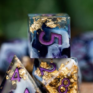 May include: A set of six polyhedral dice with a blue, white, and purple marbled design. The dice are embedded with gold flakes and have purple numbers.