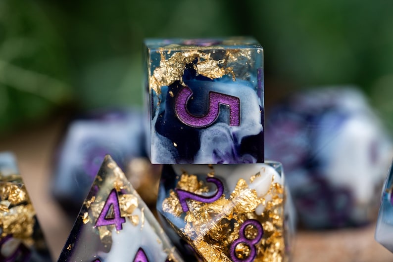 May include: A set of six polyhedral dice with a blue, white, and purple marbled design. The dice are embedded with gold flakes and have purple numbers.