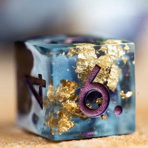 May include: A blue, clear resin dice with gold flakes and purple numbers. The dice shows the number six.