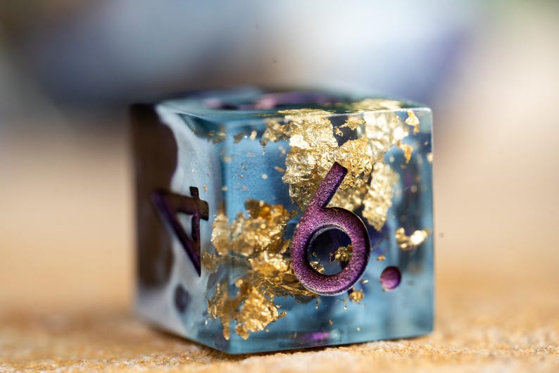May include: A blue, clear resin dice with gold flakes and purple numbers. The dice shows the number six.