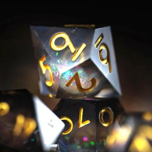 May include: Close-up of several polyhedral dice. The dice are clear with gold-colored numbers and glitter inside. The numbers are 1, 5, 6, and 9. The dice are stacked on top of each other.