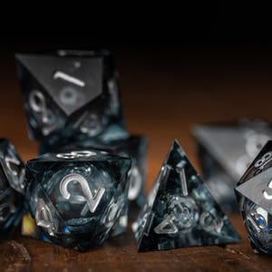 May include: A set of polyhedral dice with a dark, marbled effect. The dice are black with silver numbering, displayed on a wooden surface. The set includes various shapes, such as d4, d6, d8, d10, d12, and d20.
