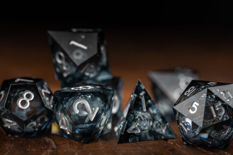 May include: A set of polyhedral dice with a dark, marbled effect. The dice are black with silver numbering, displayed on a wooden surface. The set includes various shapes, such as d4, d6, d8, d10, d12, and d20.