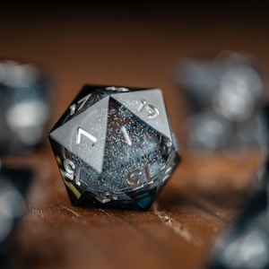 May include: A close-up shot of a black and silver twenty-sided die, with visible numbers. The die has a glittery interior and is set against a blurred background of other dice. These dice are likely used for tabletop role-playing games.