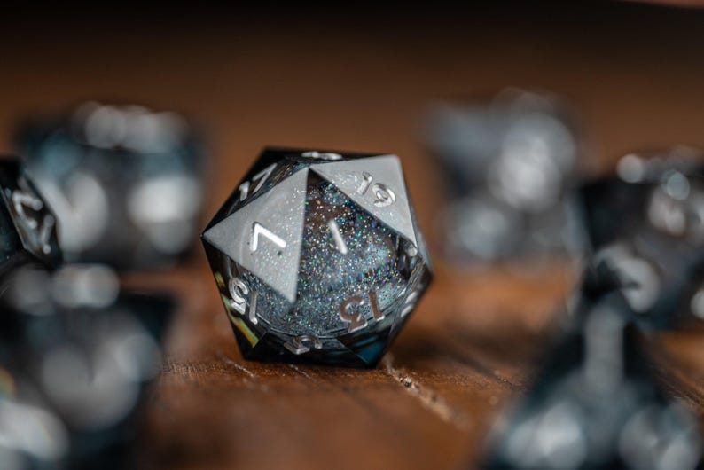 May include: A close-up shot of a black and silver twenty-sided die, with visible numbers. The die has a glittery interior and is set against a blurred background of other dice. These dice are likely used for tabletop role-playing games.