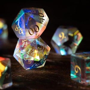 May include: Close-up of several iridescent polyhedral dice. The dice are clear with gold numbering and reflect a rainbow of colors. The dice are arranged on a dark surface, with some in focus and others blurred.
